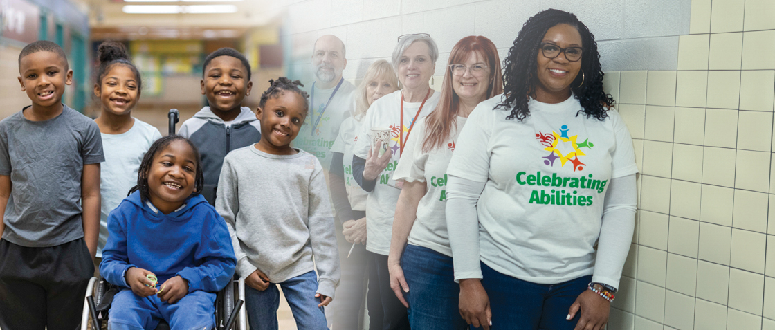 Elementary school children from diverse races and abilities along with diverse teachers smiling and wearing shirts that say "Celebrating Abilities" in the school hallway.